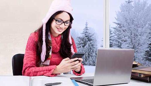 Female student studying at home while wearing sweater and using mobile phone with laptop on the table